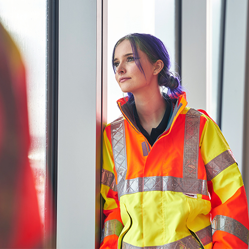 a photo of a teenage woman apprentice in a high vis jacket gazing out of an airport window