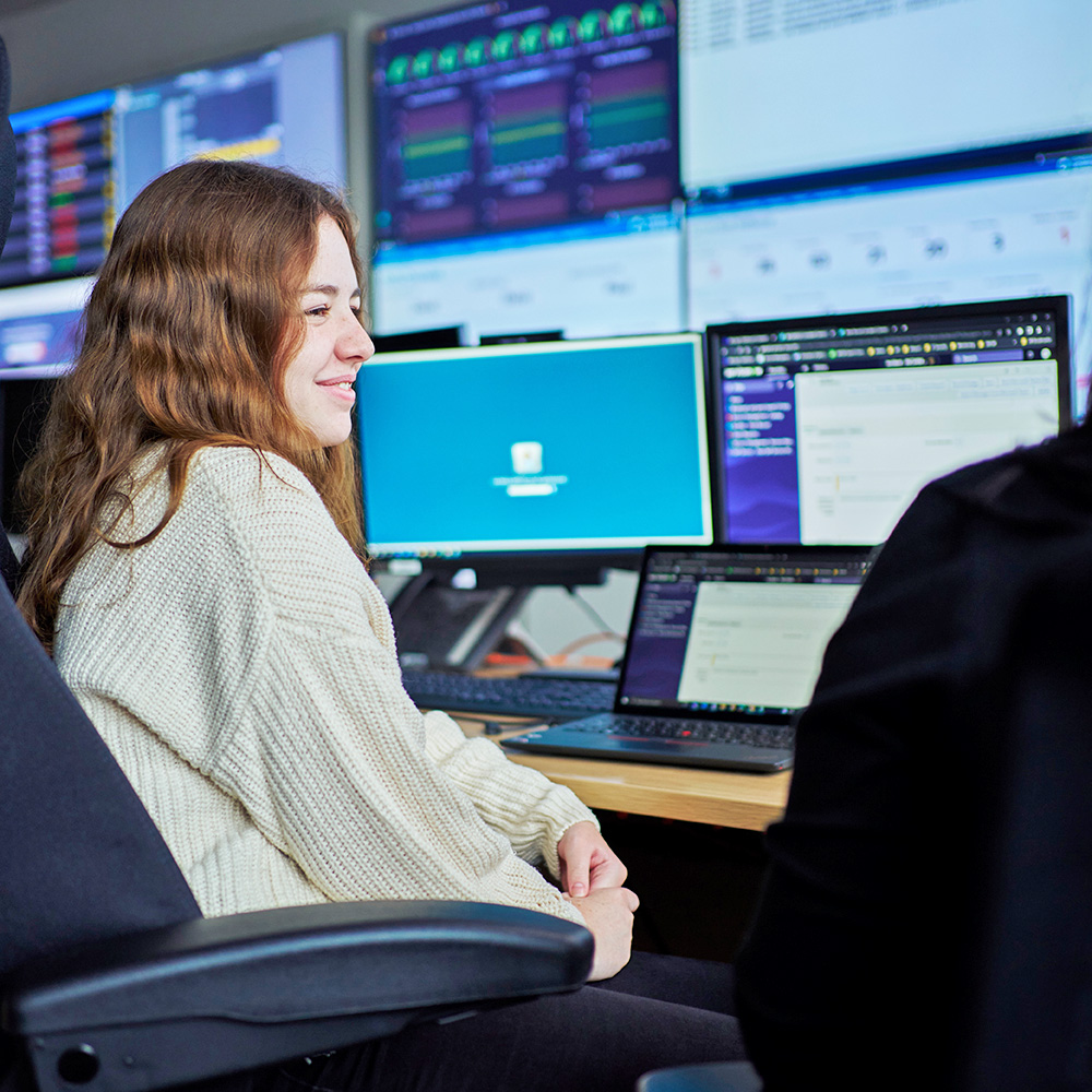 young girl sat in front of a wall of computer monitors displaying technical information