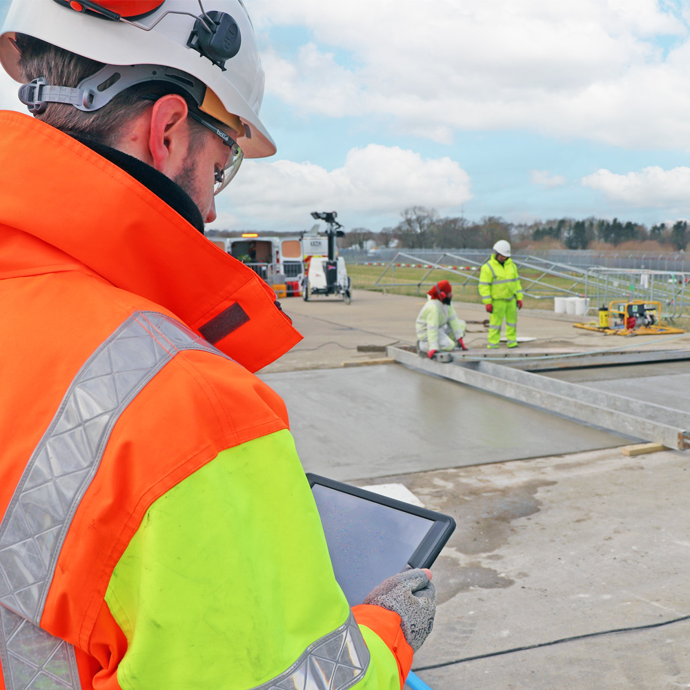 Man in high vis jacket looking at tablet with concrete laying happening in background