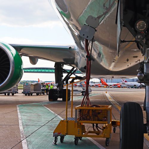 a photo from underneath a parked plane of a ground electrical power cable plugged into the fuselage, with the London Gatwick pier 6 bridge visible in the background