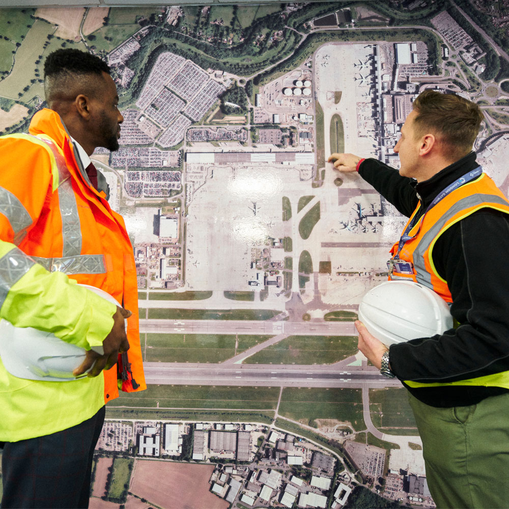 two people weith high visibility bests and hard hats under their arm talking over a wall sized aerial photograph of the airfield